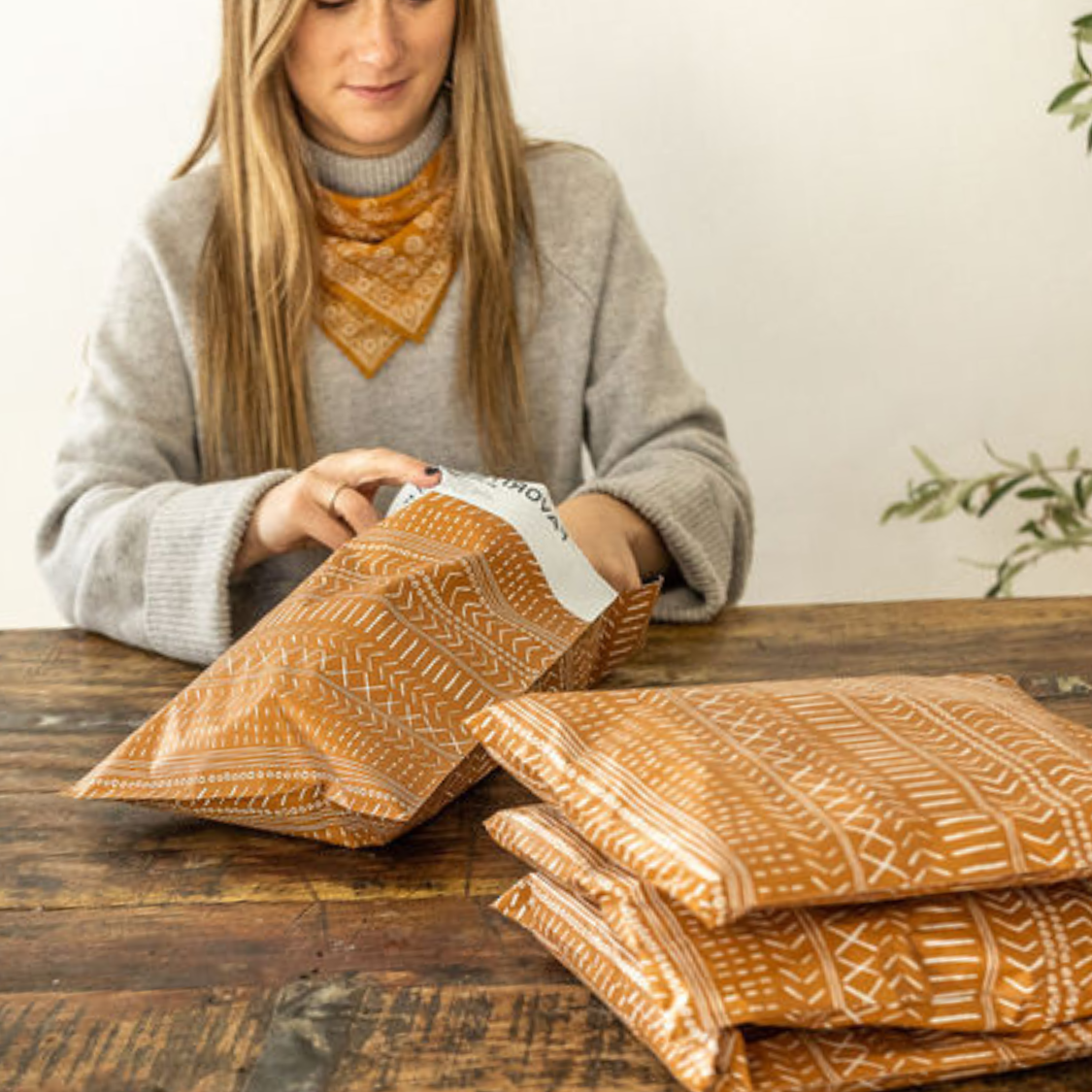 Woman packaging small business orders with Favorite Supplies' Boho Brown Poly Mailers on a table stacked with stylish shipping envelopes. Elevate your packaging game with eco-friendly, self-sealing bags.