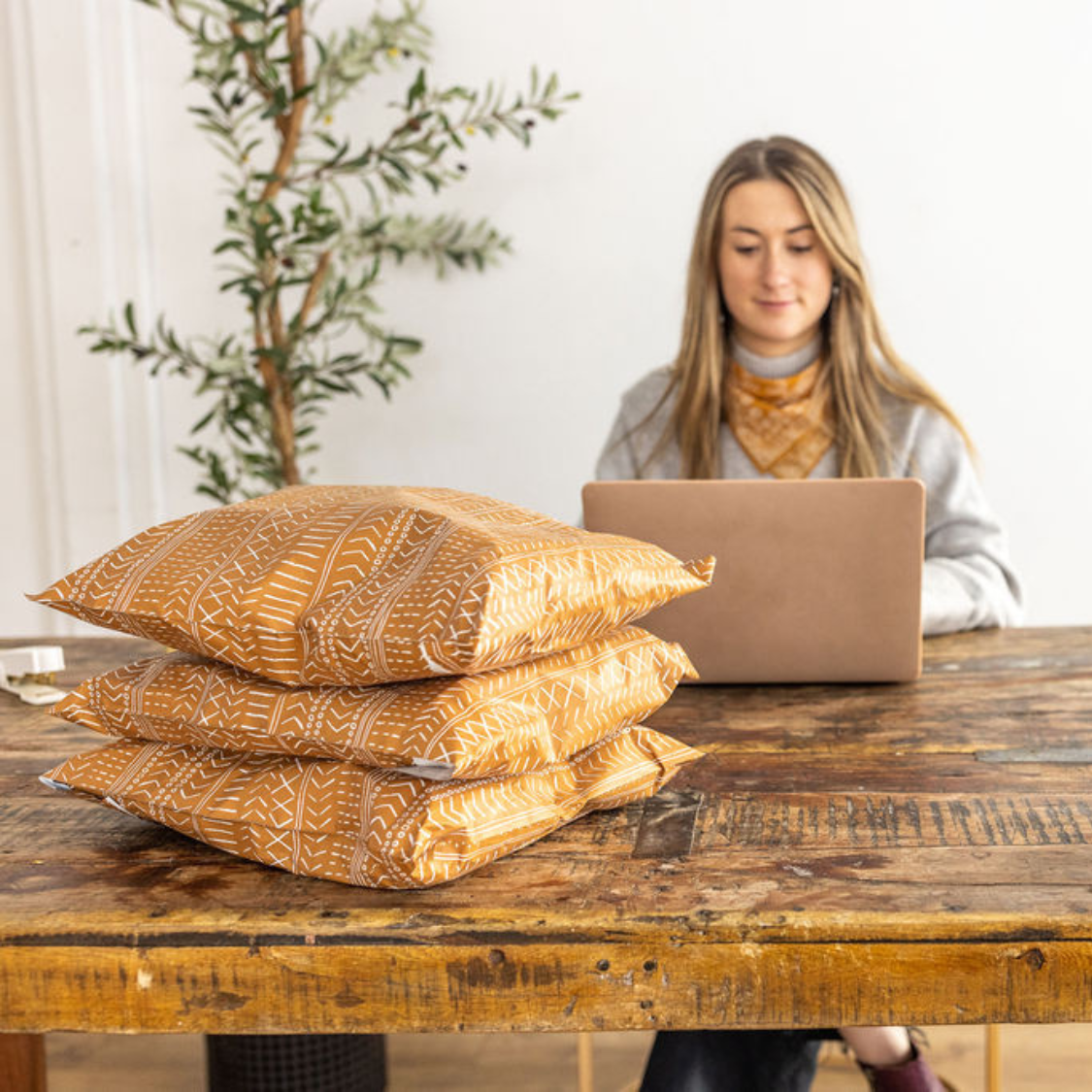 Woman processing small business orders on a laptop with a stack of Favorite Supplies' Boho Brown Poly Mailers on the table. Streamlining shipping for efficient and stylish packaging.