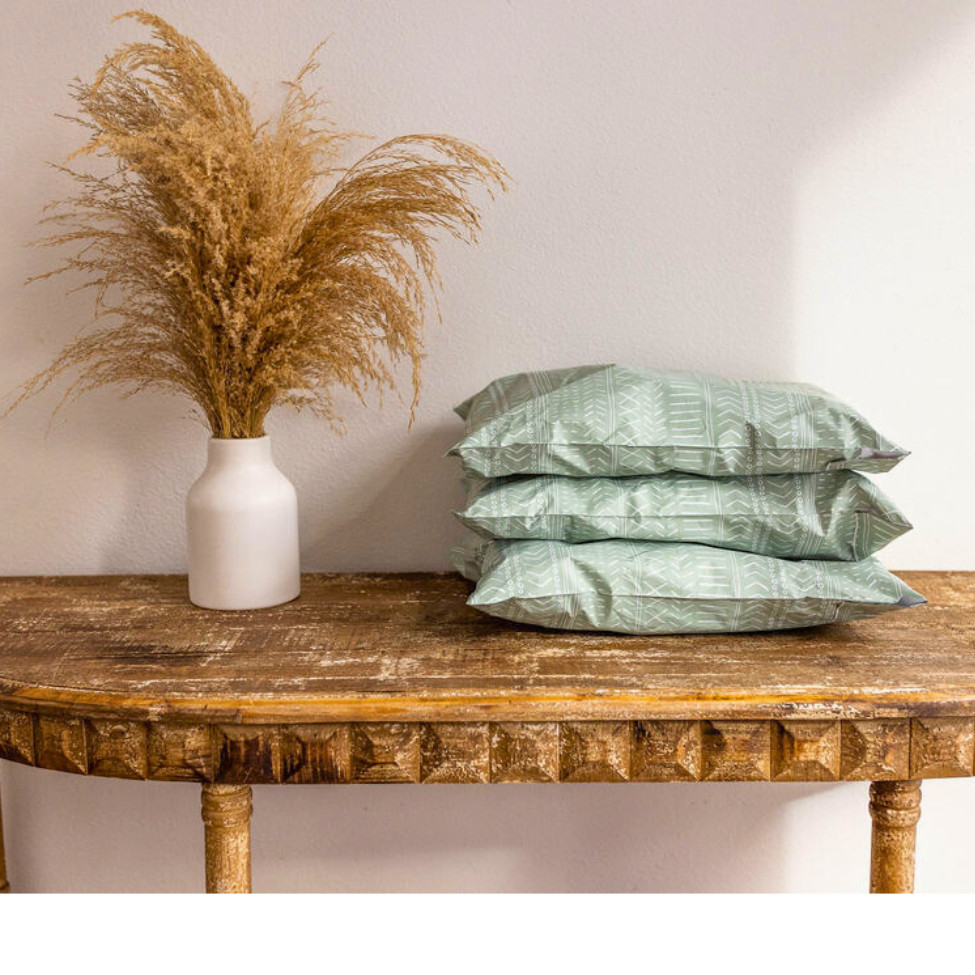 10x13 Green Geo Boho Poly Mailers stacked on a table against the wall, accompanied by a wheat plant vase. A stylish arrangement for small business shipping.