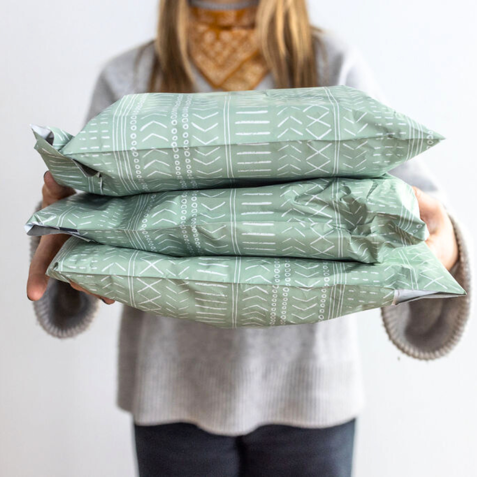 Woman against a wall, holding three 10x13 Green Geo Boho Poly Mailers in a close-up. Highlighting the charming pattern and the convenient size for small business shipping.