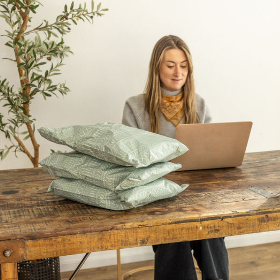 Woman on laptop with 10x13 Green Geo Boho Poly Mailers on the table. Soft lighting enhances the stylish design for convenient small business shipping.