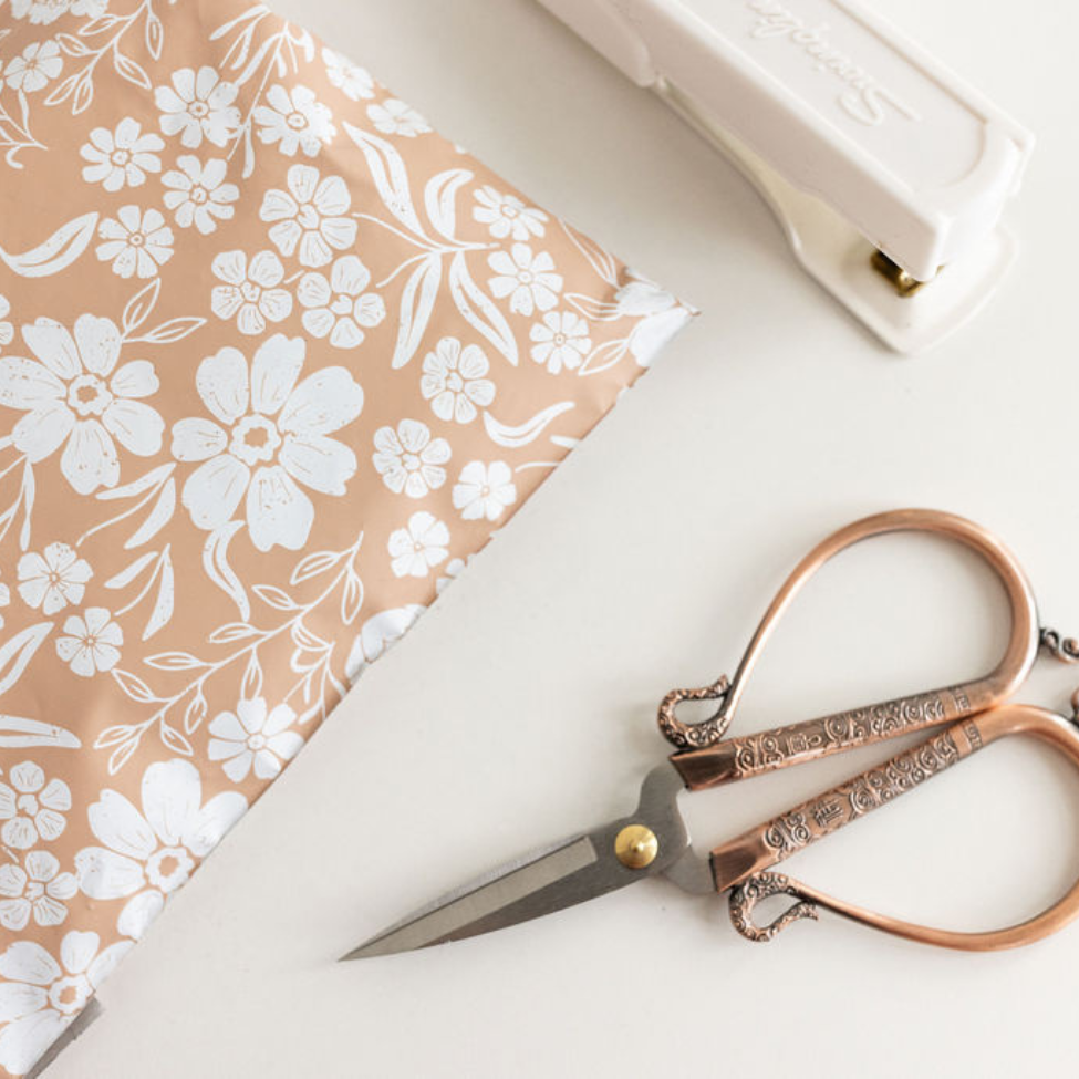 Floral Dusty Pink Poly Mailer on a white table, accompanied by old-fashioned scissors and a stapler. A charming scene showcasing stylish packaging tools by Favorite Supplies.