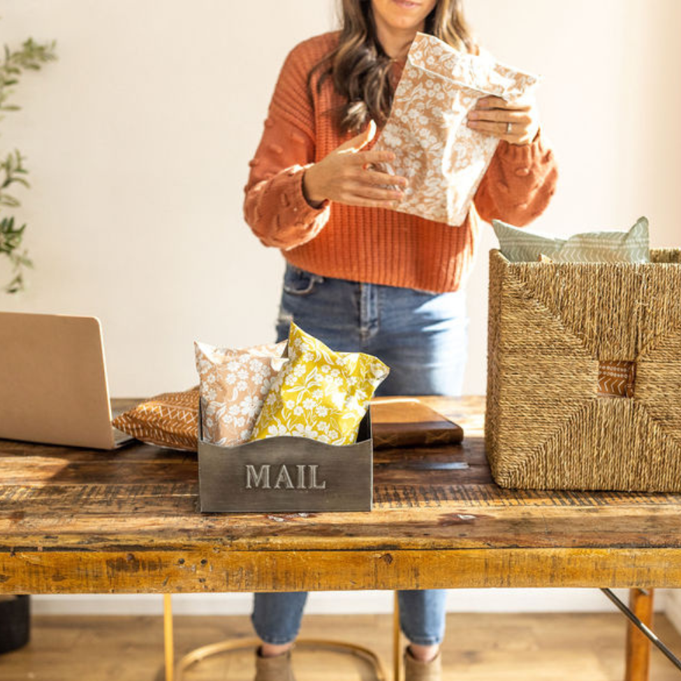 Woman shipping and packaging with various colored mailers, including Floral Dusty Pink. Laptop and mail basket in hand, highlighting a stylish and organized shipping process by Favorite Supplies.