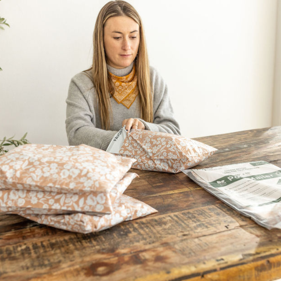 Woman packing with an unopened 10x13 Floral Dusty Pink Poly Mailer package nearby. Finalizing mailers with a stack on the side, showcasing the stylish packaging process by Favorite Supplies.