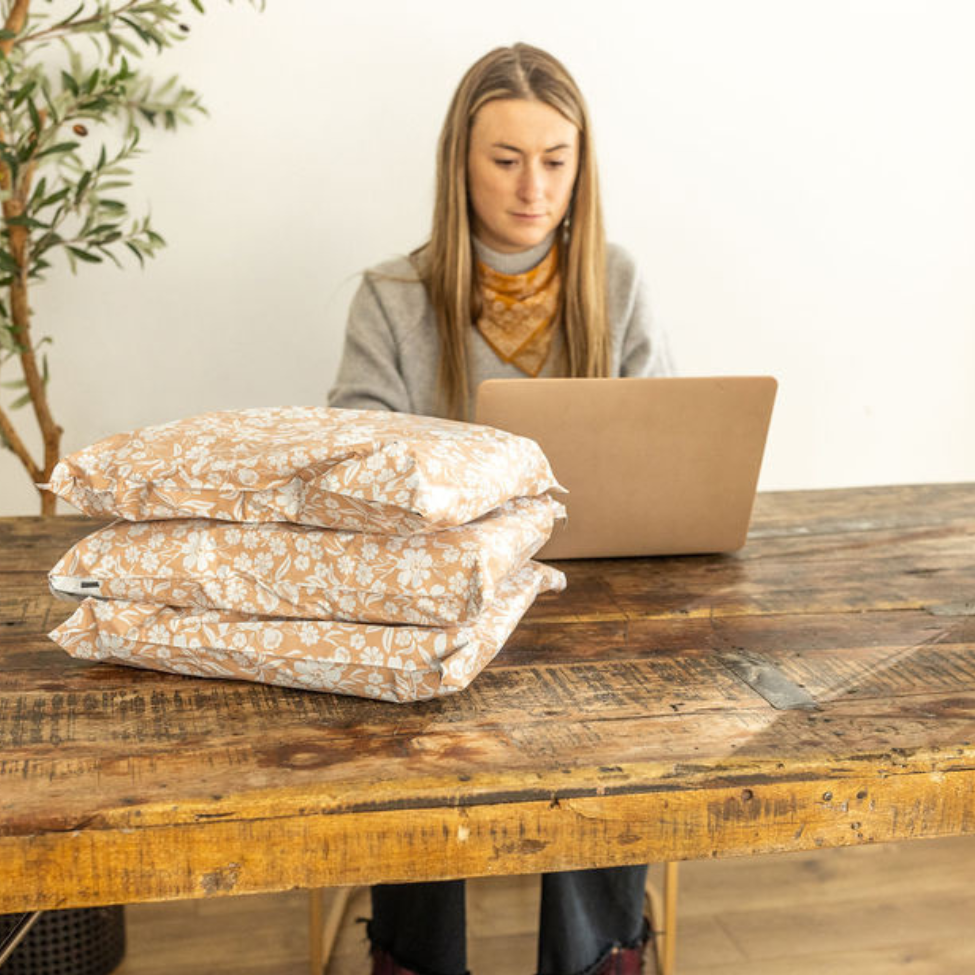 Woman working on her computer with a stack of 10x13 Floral Dusty Pink Poly Mailers on the table. Ready for shipping, highlighting the efficiency and style in small business packaging by Favorite Supplies.